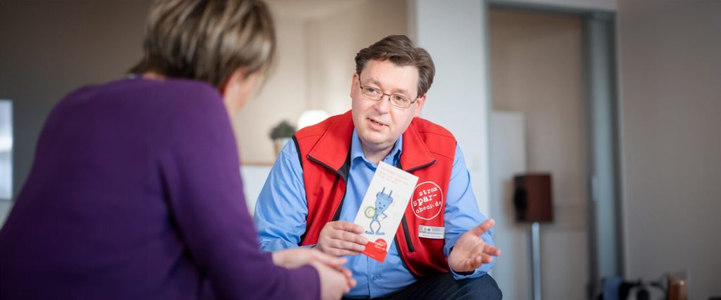 Ein Stromspar-Helfer in roter Weste berät eine Person in deren Wohnung. Sie sitzen zusammen und sprechen über den Energiesparen.